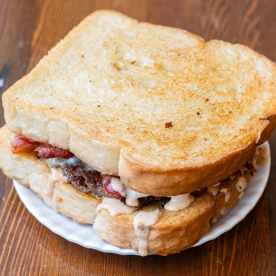 Iggy's Eggies sandwich with toasted bread, bacon, patty, and creamy sauce on a white plate.