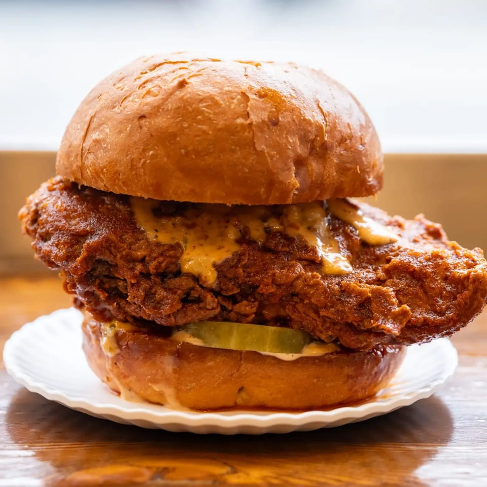 Crispy fried chicken sandwich with sauce and pickles on a brioche bun, served on a white plate.