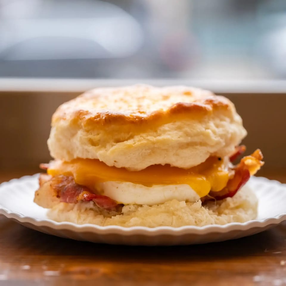 Golden-brown biscuit sandwich with bacon, egg, and melted cheese on a white plate.