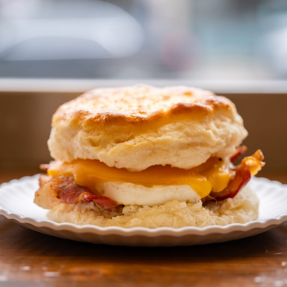 A fluffy biscuit sandwich with bacon, egg, and melted cheese on a white plate.